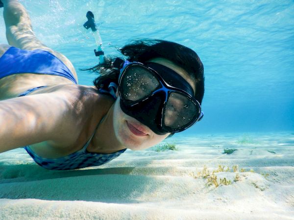 A person is snorkeling underwater, wearing a blue swimsuit and snorkel mask, swimming above sandy ocean floor with clear blue water.