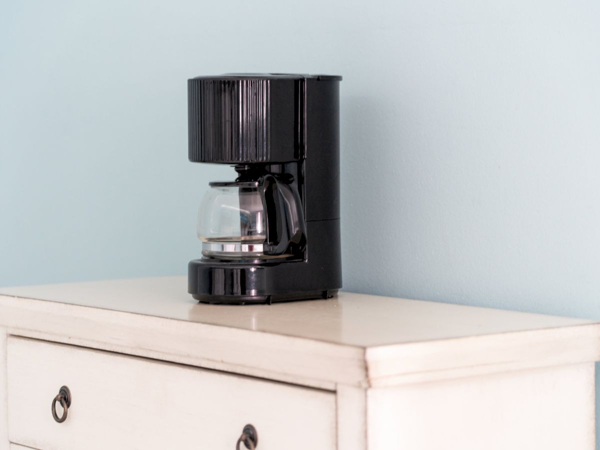 A black drip coffee machine with a glass carafe sits on a light cabinet shelf in a minimal room.