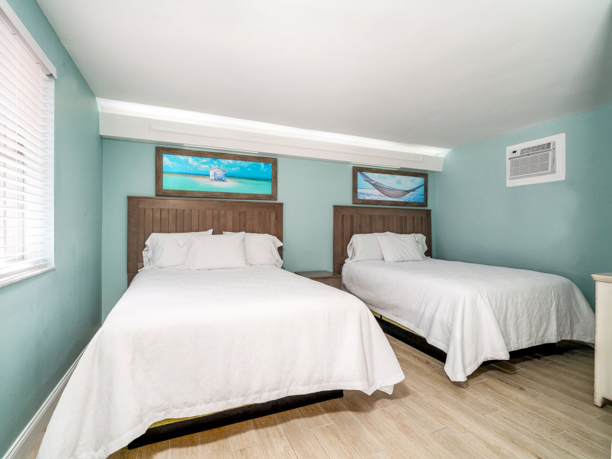 Bright bedroom with two twin beds, teal walls, beach-themed framed art, and a wall AC unit, with blinds on the window.