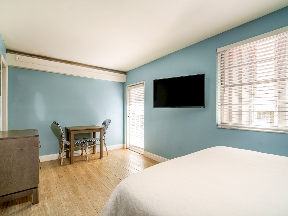 A small living room with blue walls, a wall-mounted TV, two windows with blinds, and a table with patterned chairs.
