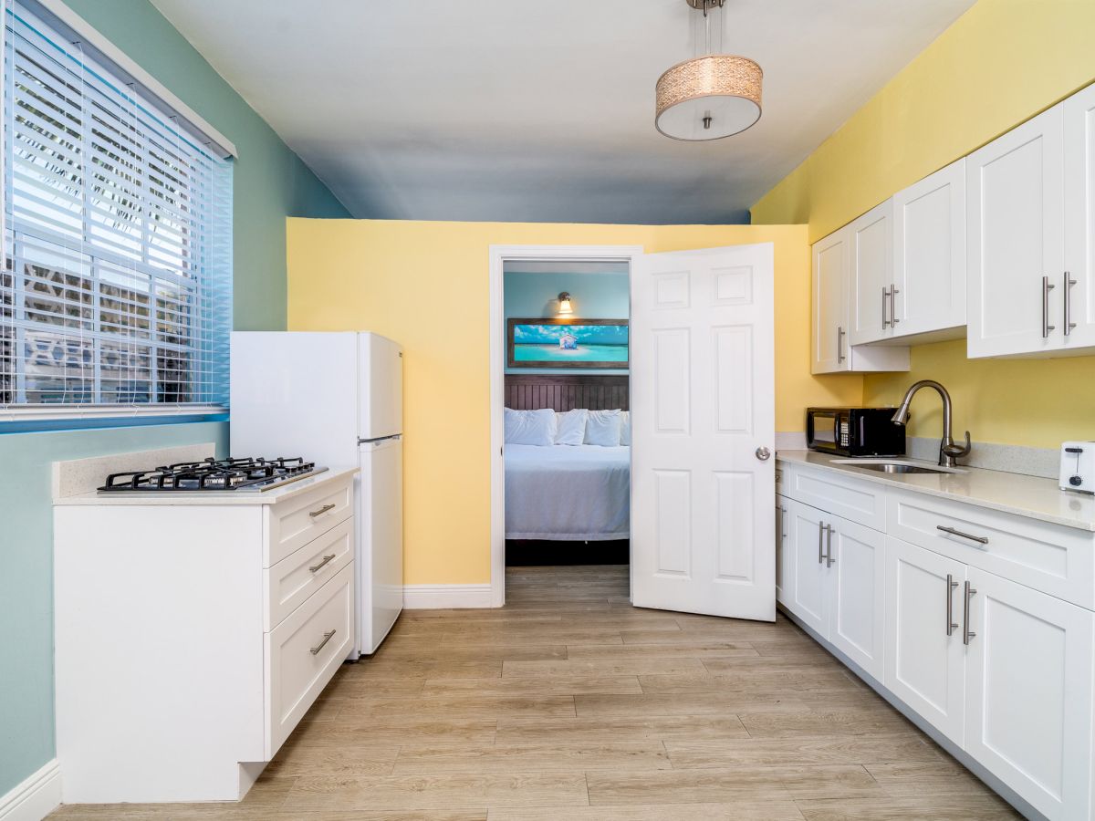 A bright modern kitchen with white cabinets, granite counters, gas stove, sink and coffee maker, and a doorway to the bedroom.