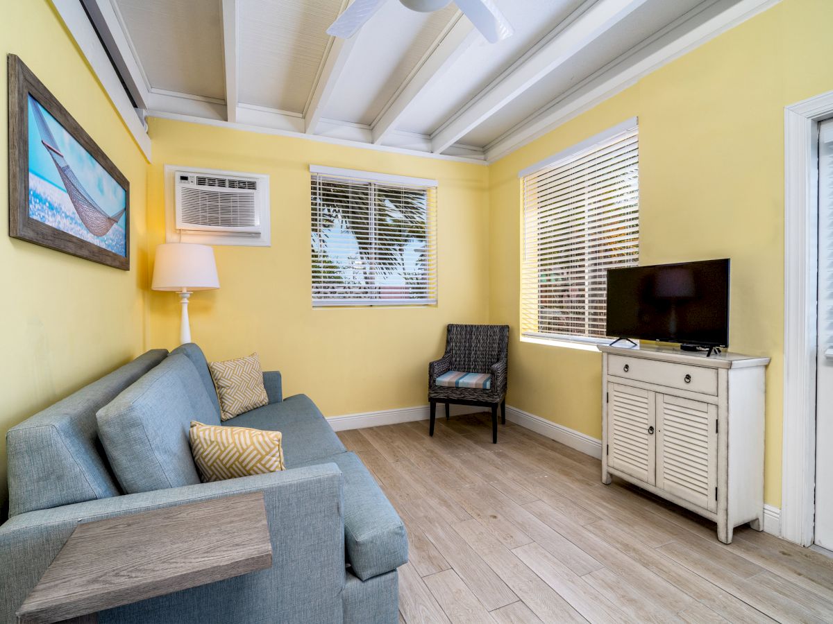 Bright living room with yellow walls, a sofa, a TV on a cabinet, ceiling beams, an air conditioner, and three windows with blinds.
