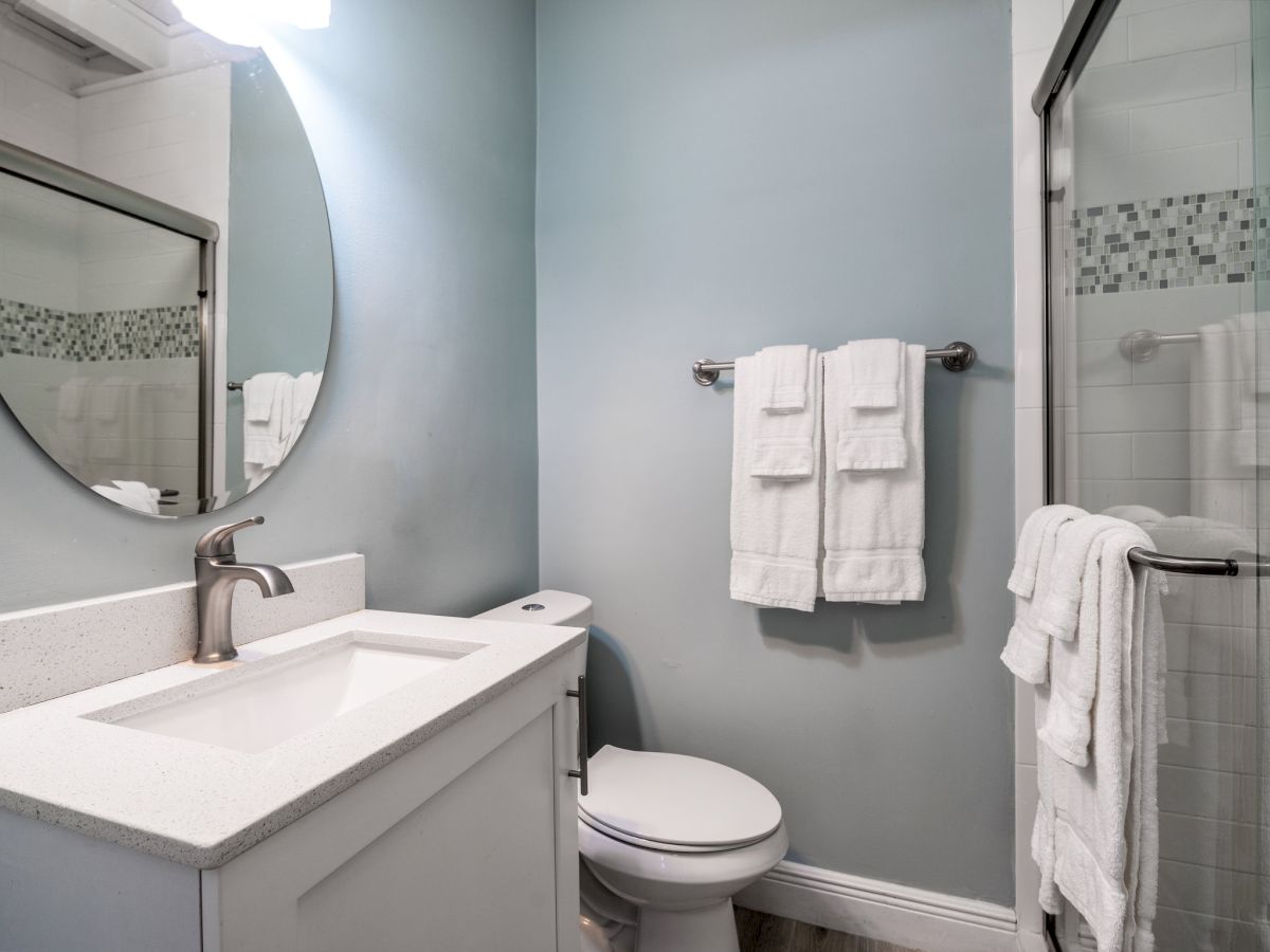 A bright bathroom with a sink and mirror, a toilet, and neatly hung white towels beside a glass shower.