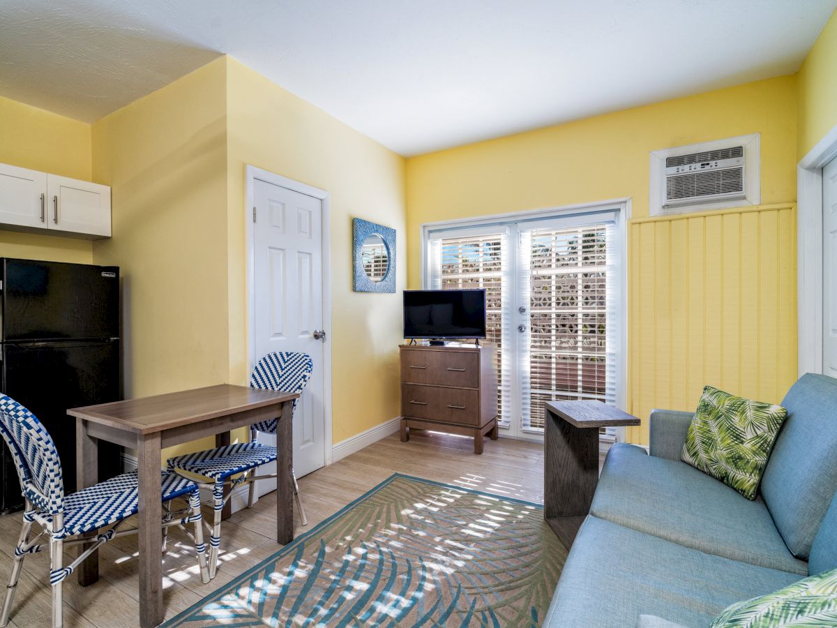 A bright yellow living room with a kitchenette, dining table, sofa, TV on a cabinet, windows with blinds, and a wall air conditioner.