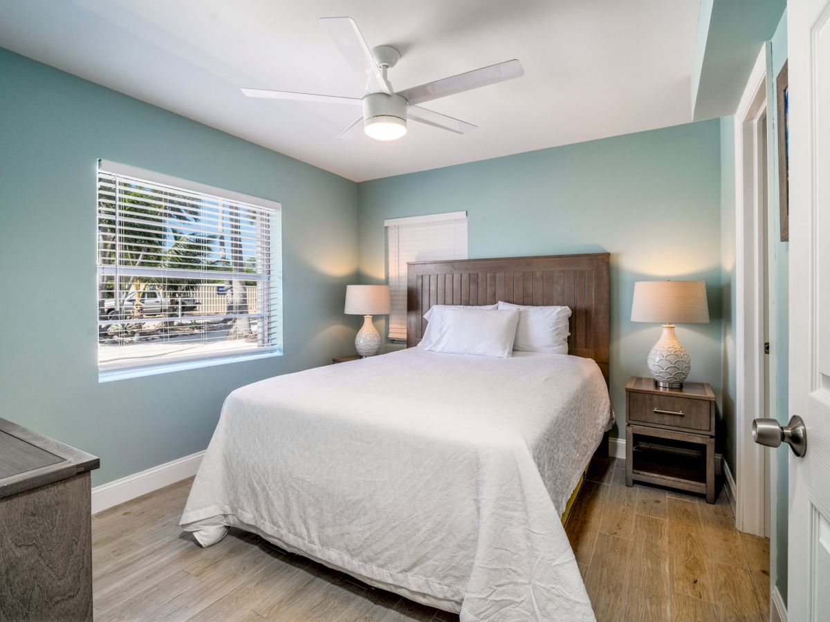 Bright modern bedroom with a white bed, wood headboard, bedside lamps, blinds, ceiling fan, and teal walls.