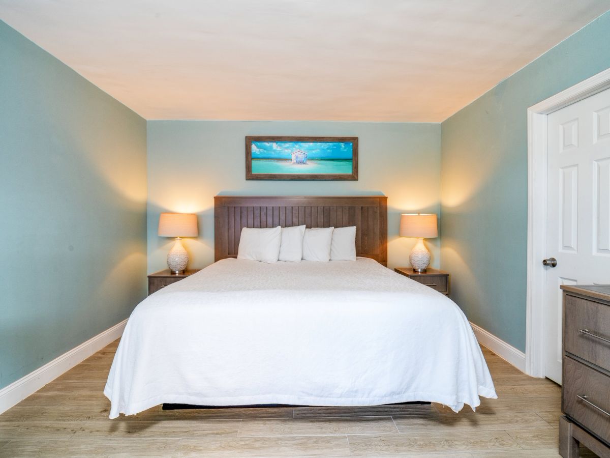 A tidy bedroom with a large bed, two bedside tables and lamps, a beach wall painting, and a white closet door.