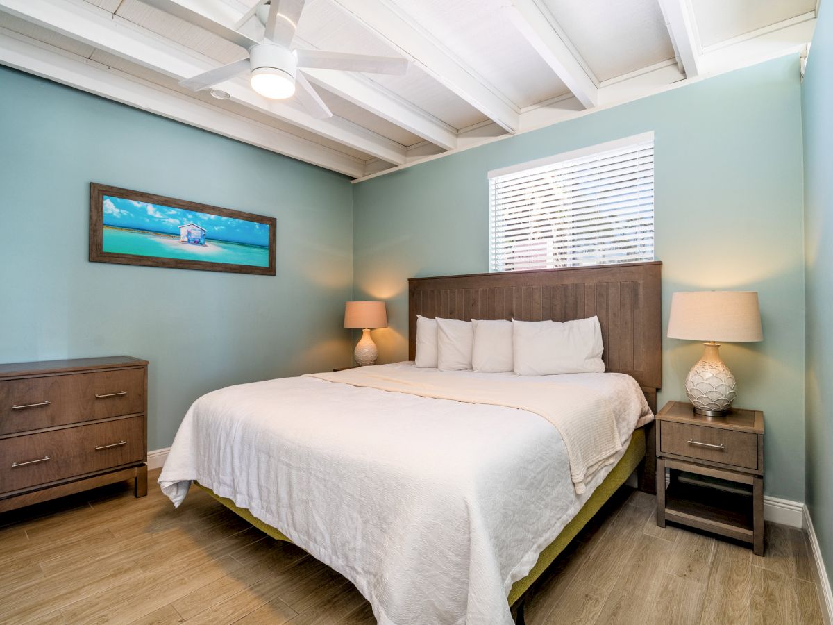 A bright modern bedroom with a wooden bed, nightstands, lamps, framed beach art, and window blinds.