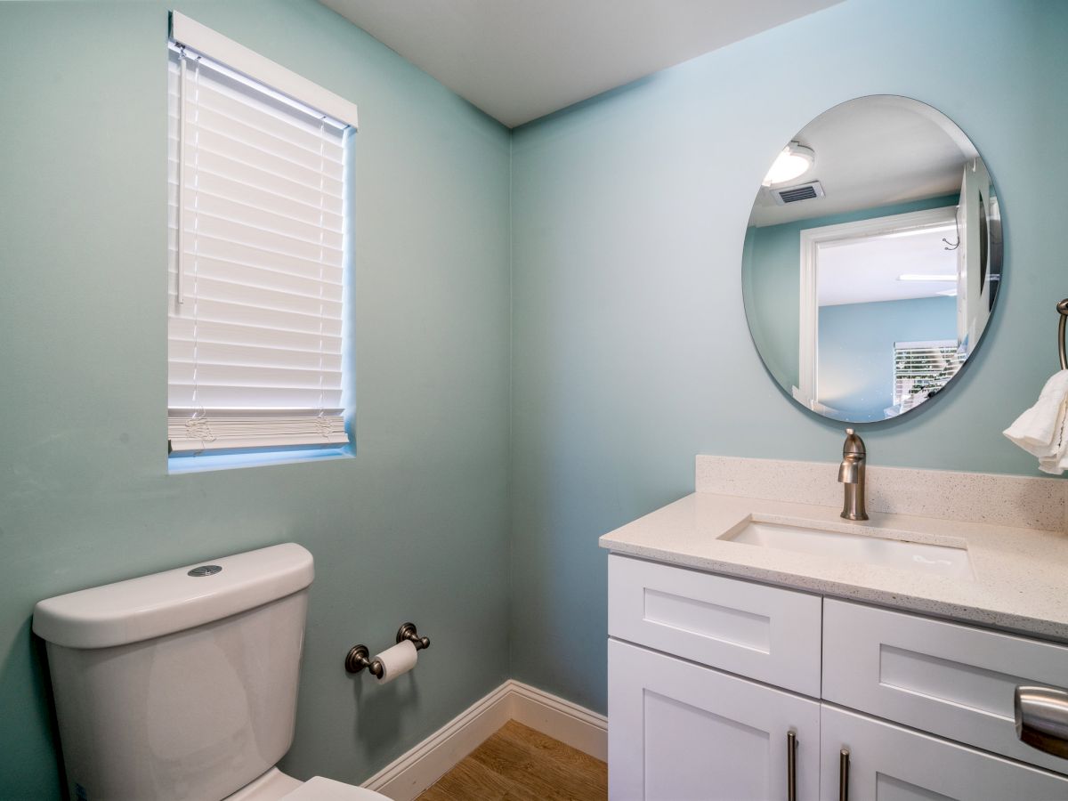Bright bathroom with teal walls, a round mirror, vanity sink, toilet, and window blinds.