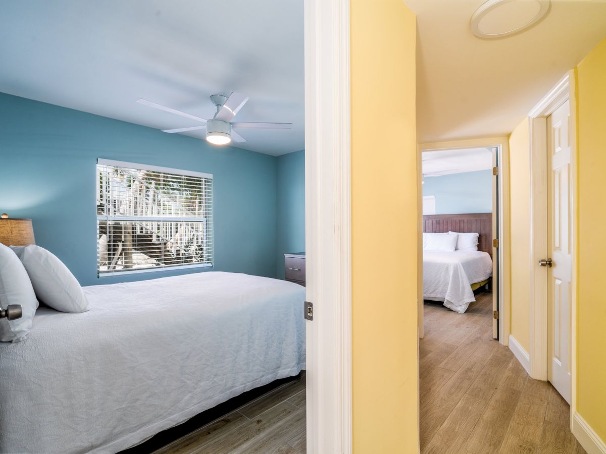 Bright bedroom with a blue accent wall, ceiling fan, window blinds, and an open door revealing another bed and room.