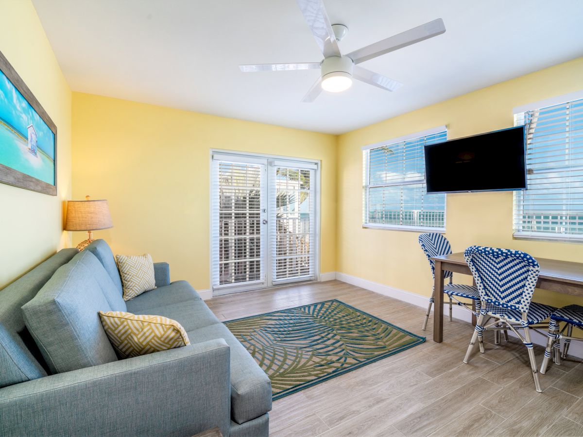 A bright living area with a ceiling fan, large TV, blue-striped chairs, windows with blinds, and an outdoor patio door.