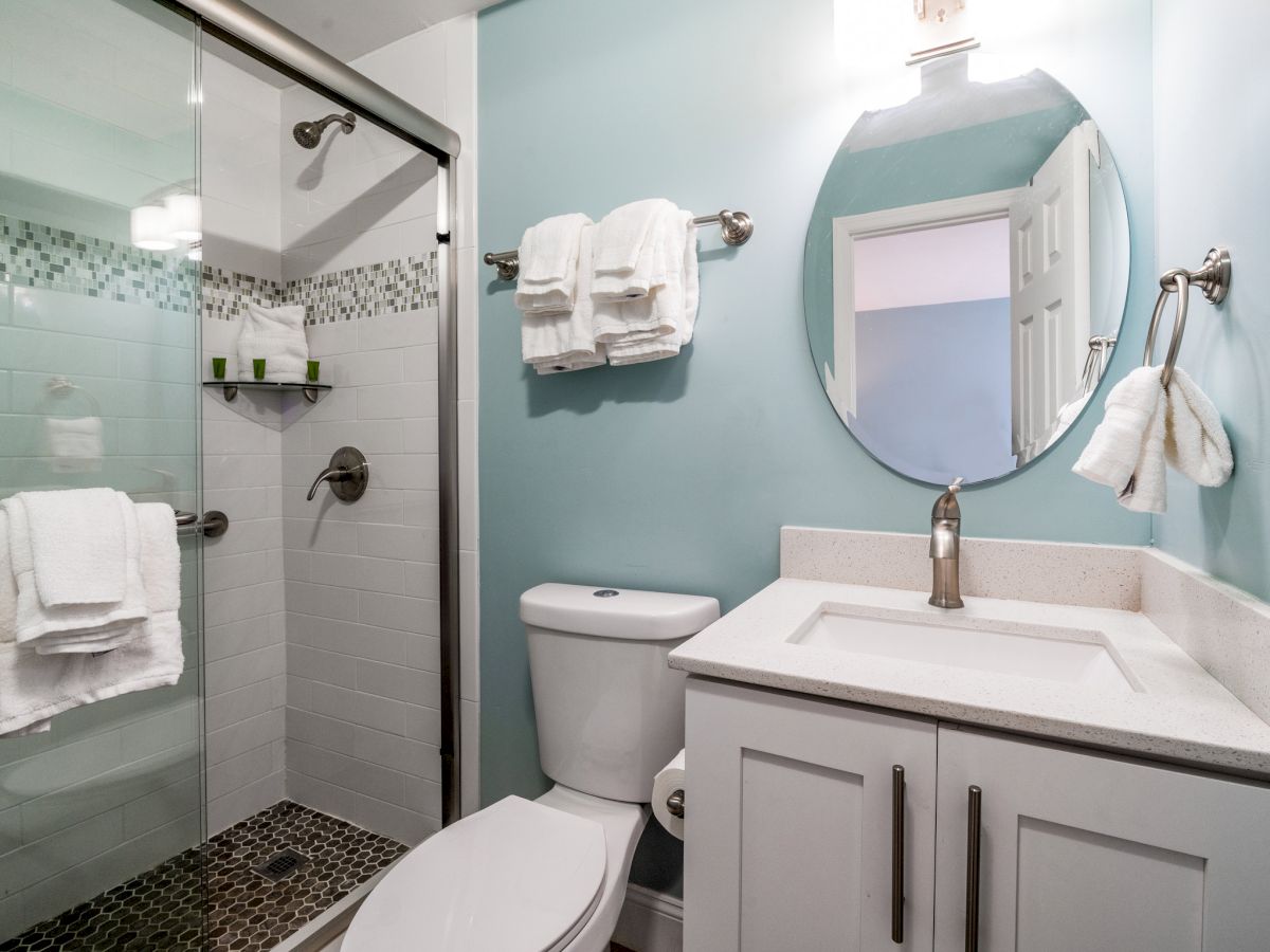 A bright bathroom with a glass shower, stacked white towels, a pedestal sink, and a large round mirror reflected in the wall.