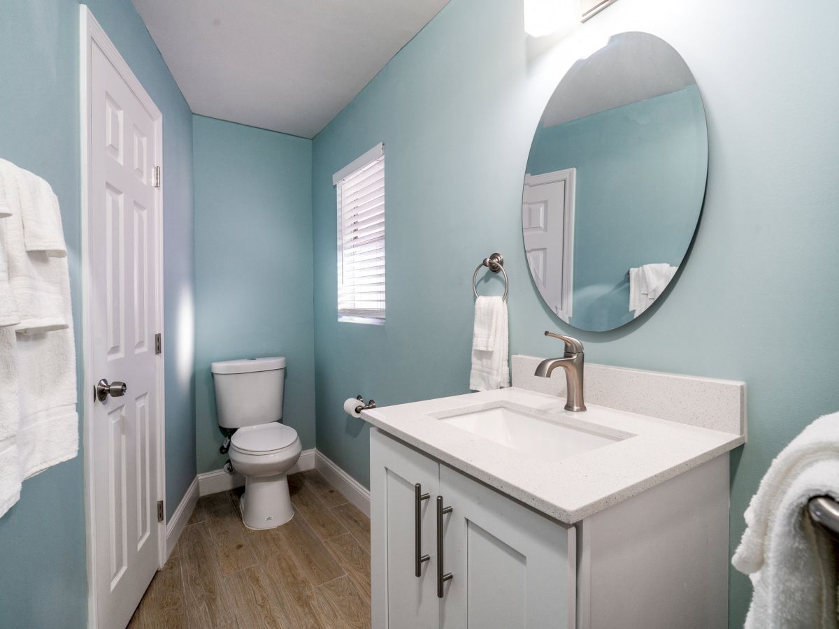 A bright bathroom with blue walls, a white vanity and sink, a large round mirror, and a toilet with towels hanging.
