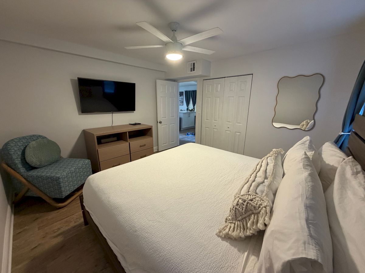 A bright bedroom with a bed, ceiling fan, wall TV, dresser, closet doors, and a decorative wavy mirror.