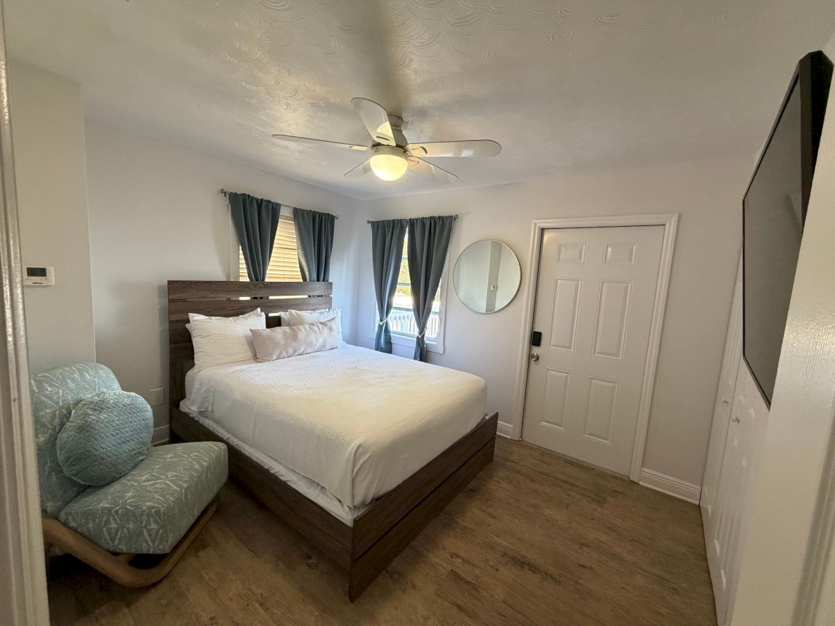 A tidy bedroom with a wooden bed, white linens, blue curtains, a ceiling fan, and a door and round wall mirror.