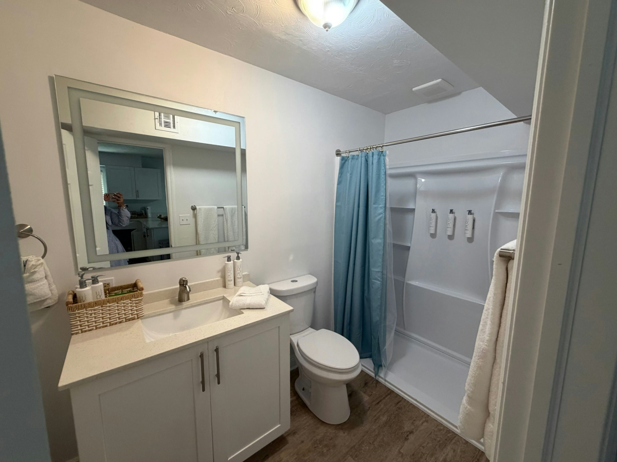 Clean modern bathroom with white vanity, large mirror, toiletries, toilet, and a blue shower curtain in the tub.