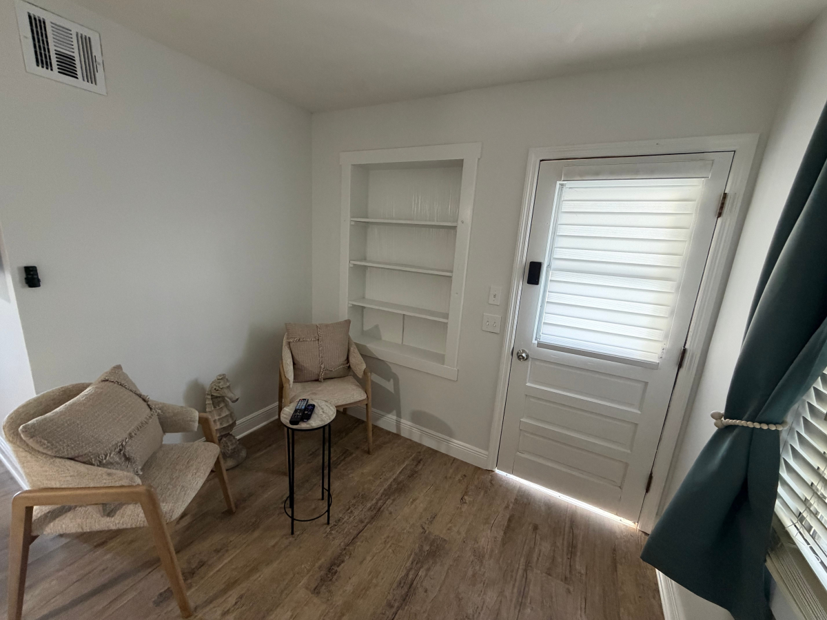 A cozy corner room with a chair and side table, open shelving, a white door with blinds, and a window curtain.