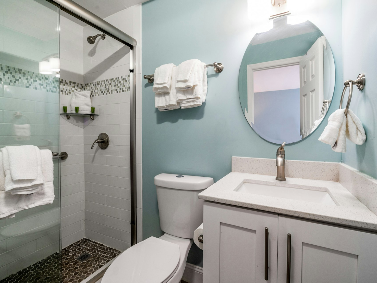 A bright bathroom with a glass shower, white towels, a round mirror, a toilet, and a countertop sink with faucet, all in calm blue tones.