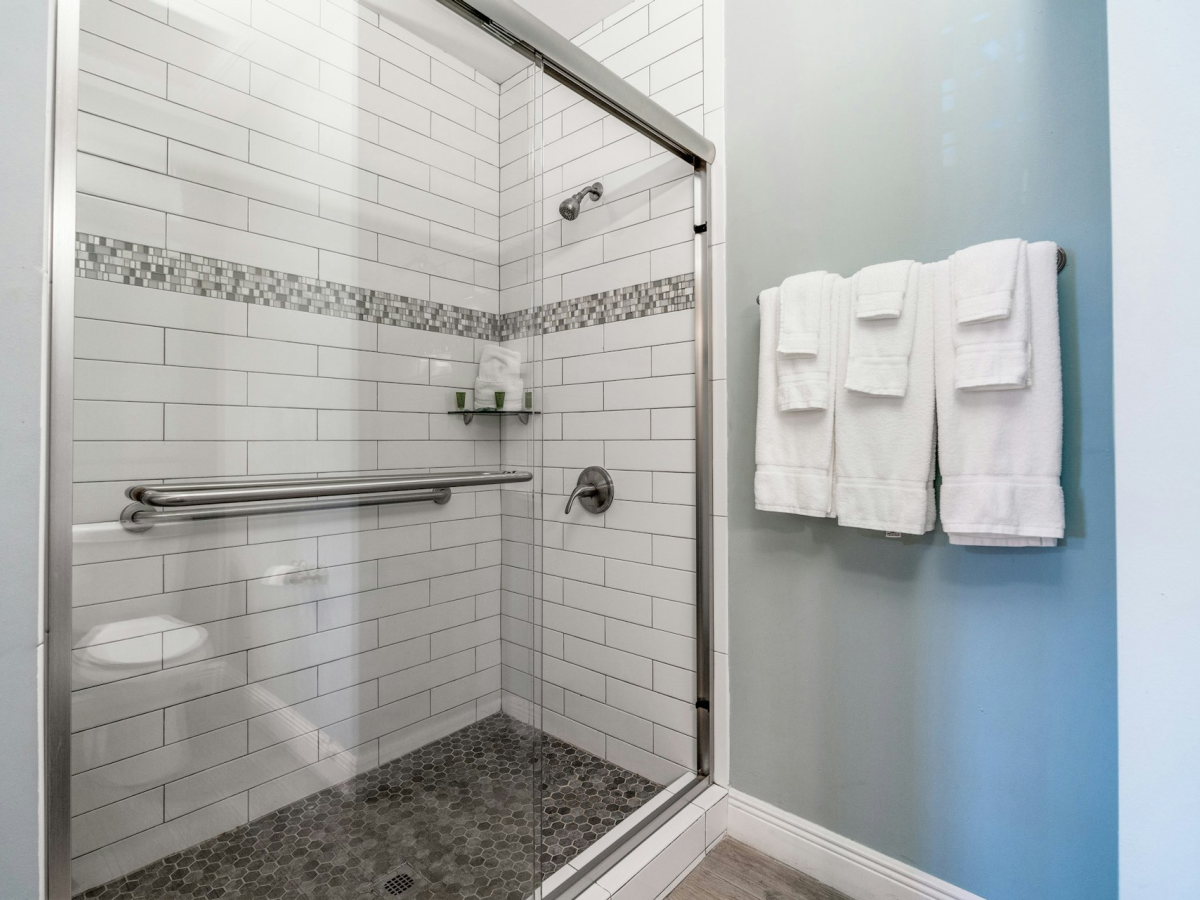 Clean tiled walk-in shower with glass door, rainfall head, grab bars, and folded white towels hanging on a blue wall.