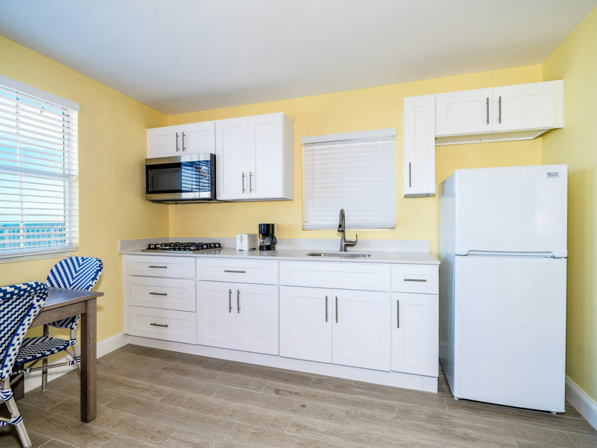 Bright kitchen with white cabinets, countertop, stove, microwave, sink, refrigerator, and blinds by two windows.