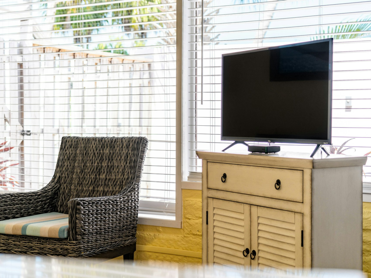 A flat-screen TV sits on a cabinet beside a wicker chair in a bright room with large windows and blinds, lit by daylight.