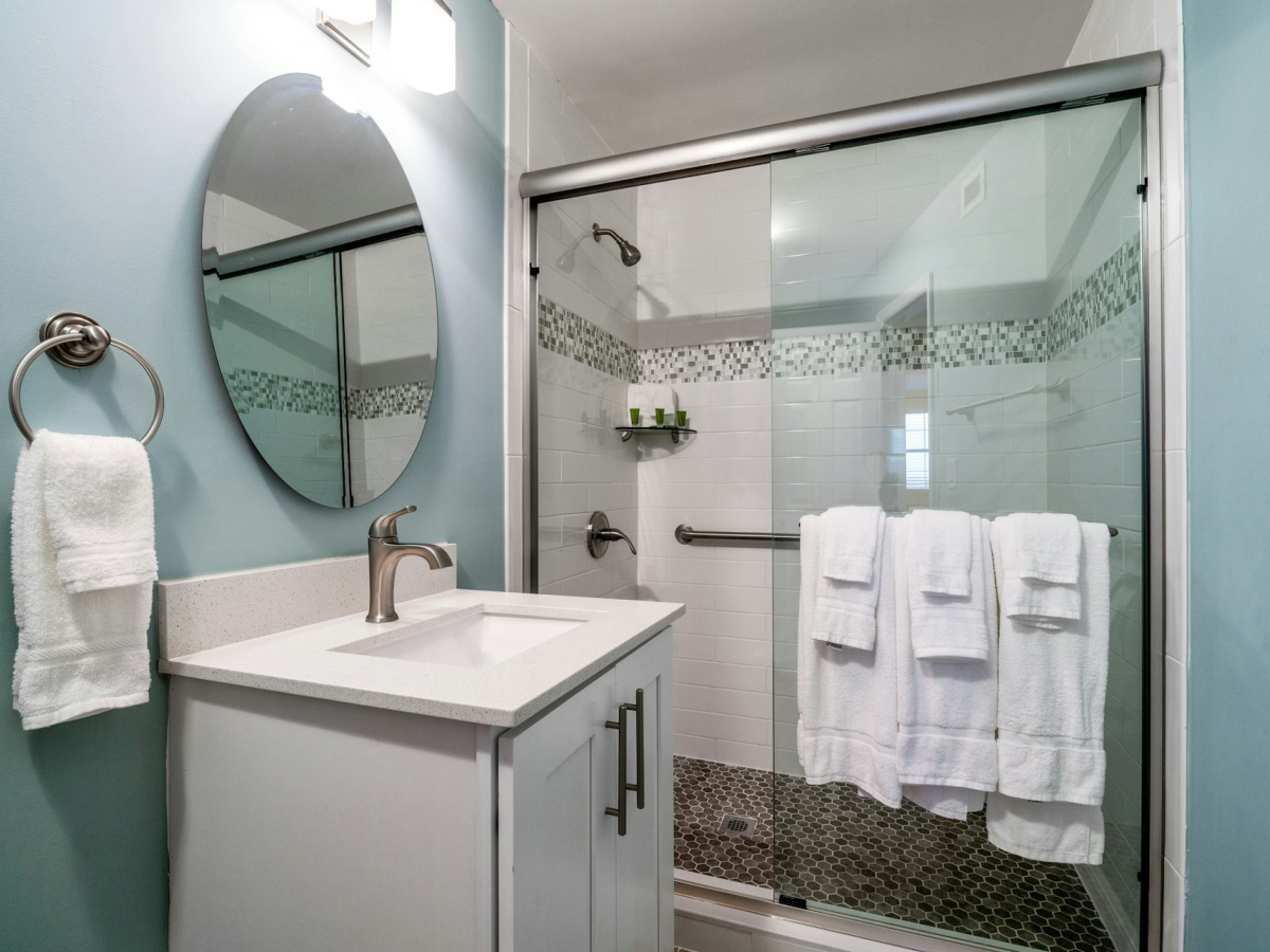 A clean modern bathroom with a white vanity sink, wall mirror, and a glass shower enclosure with neatly folded towels.