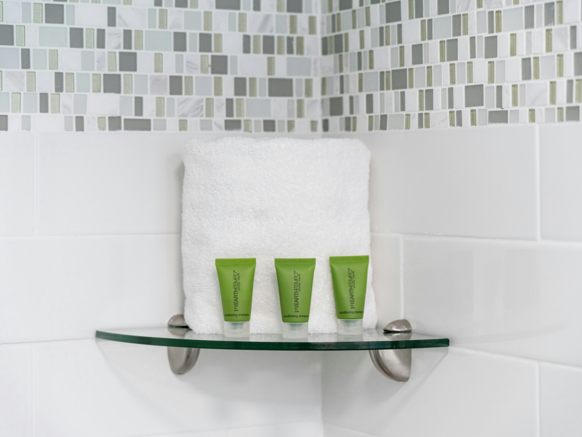 A bathroom wall shelf holding a folded white towel and three green shampoo/conditioner tubes against a tiled backsplash.