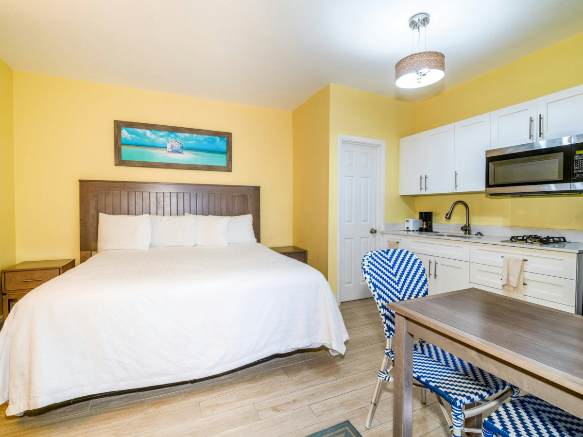 Bright studio shows a bed, white kitchenette with microwave and stove, coffee maker, and a small dining table with blue chairs.