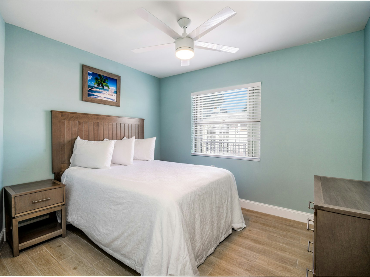 A bright bedroom with a bed, nightstands, ceiling fan light, framed beach wall art, and a window with white blinds.