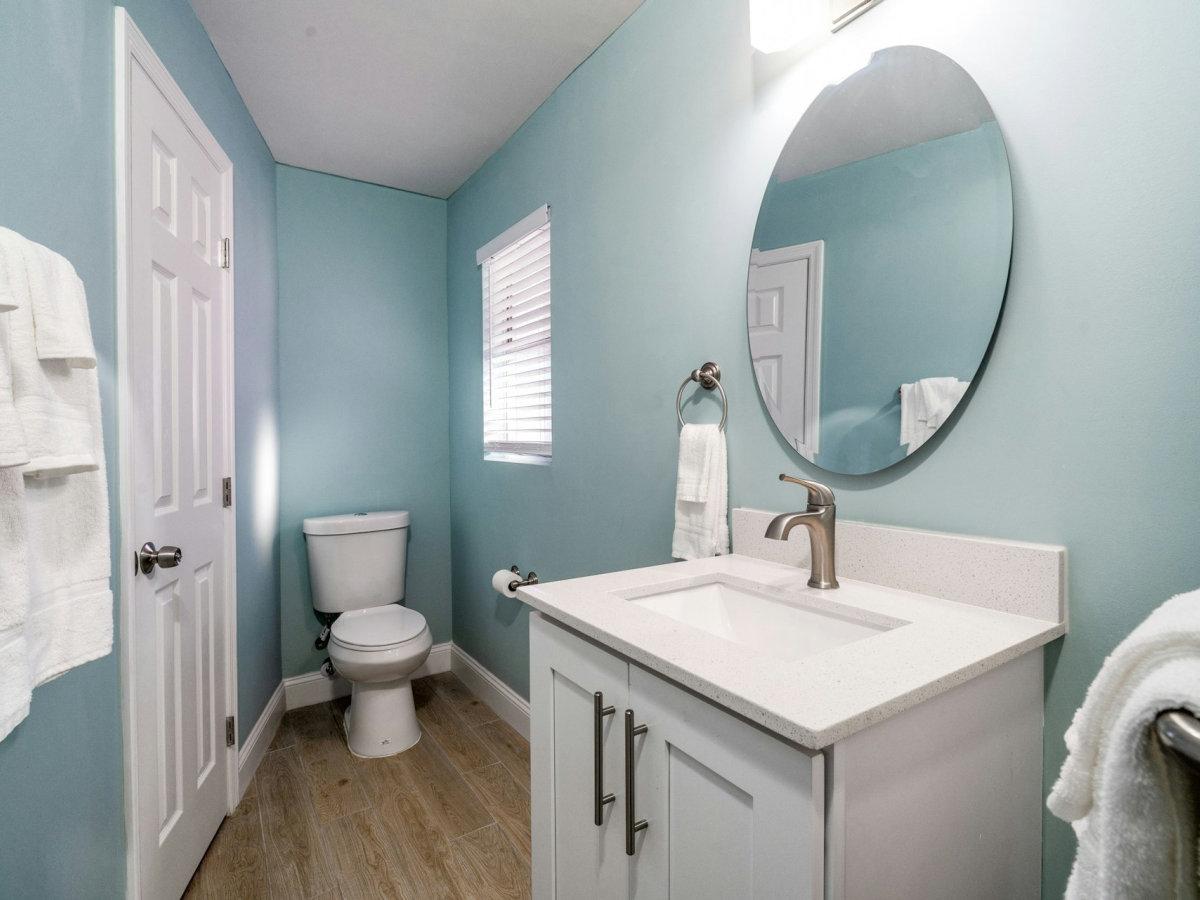 Bright bathroom with a white vanity and sink, oval mirror, toilet, and neatly folded towels on blue walls.