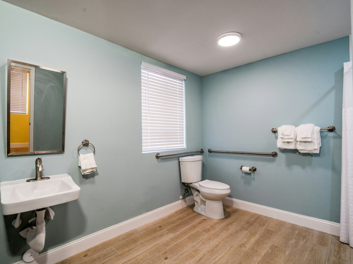 A small bathroom with a toilet, pedestal sink, towel bars, window blinds, and a ceiling light.