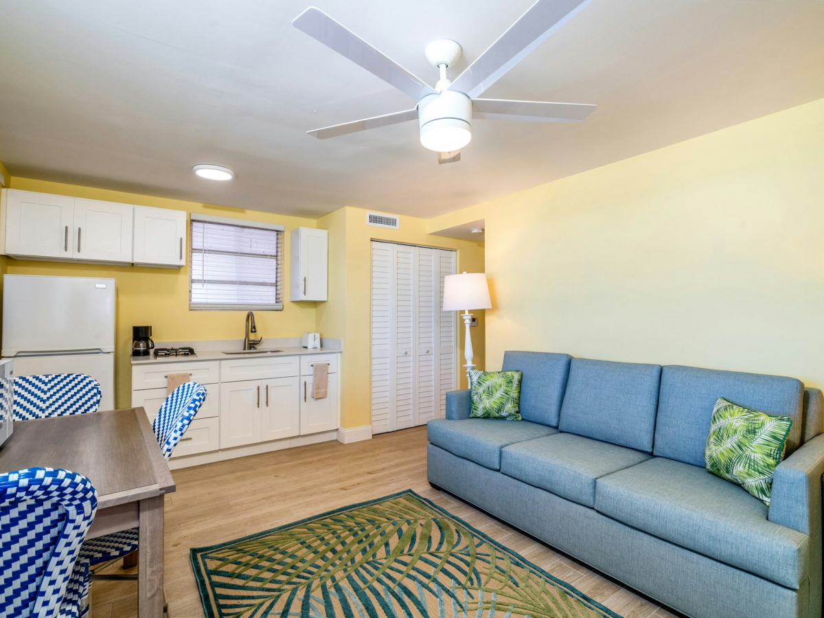 A bright living room with a blue sofa, kitchen counter and cabinets, ceiling fan with light, and a dining table with patterned cushions.