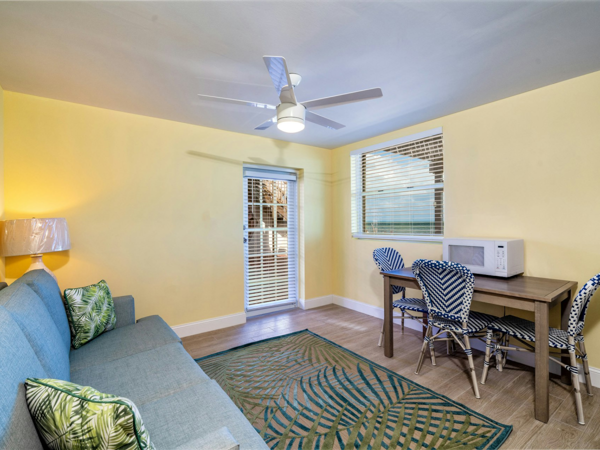 Bright living room with blue sofa, patterned rug, ceiling fan, small dining table with chairs, microwave, and window blinds.