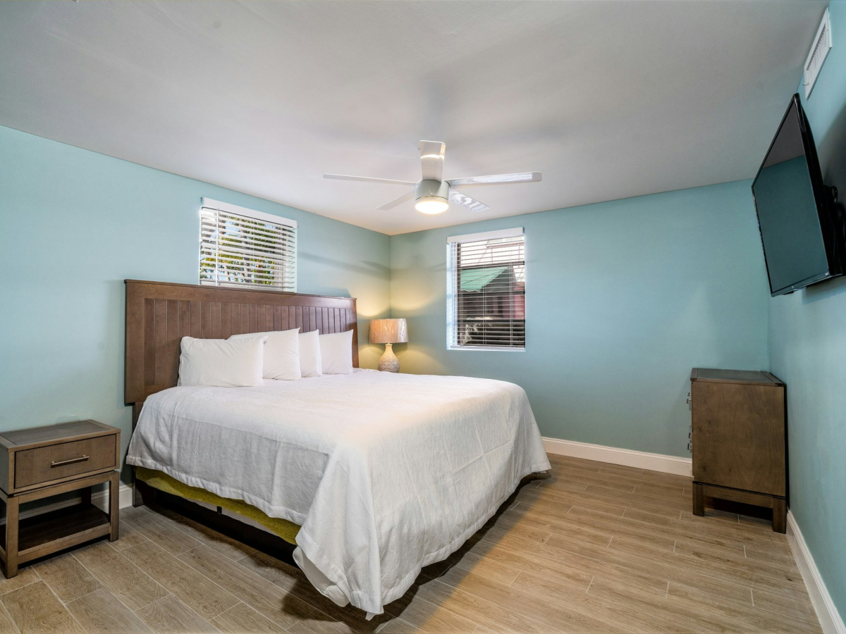 A tidy bedroom with a bed, nightstands, dresser, ceiling fan/light, windows with blinds, and a wall-mounted TV.
