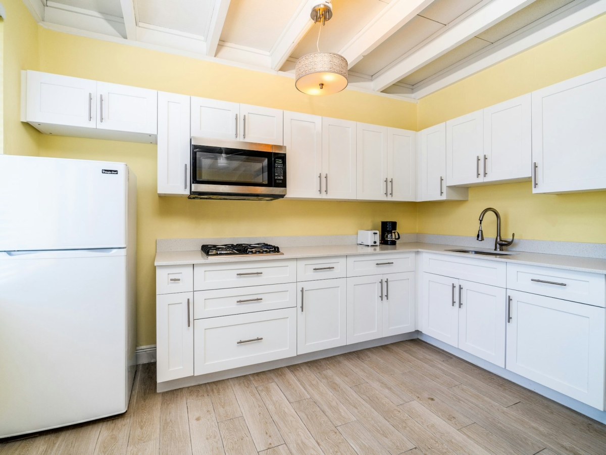 Bright kitchen with white cabinets, microwave, stove, refrigerator, coffee maker, and stainless sink with faucet.