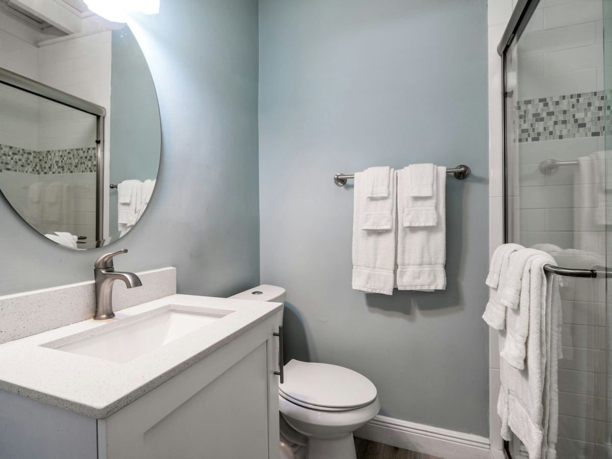 A bright bathroom with a vanity sink, wall mirror, toilet, and a glass shower with neatly folded white towels.