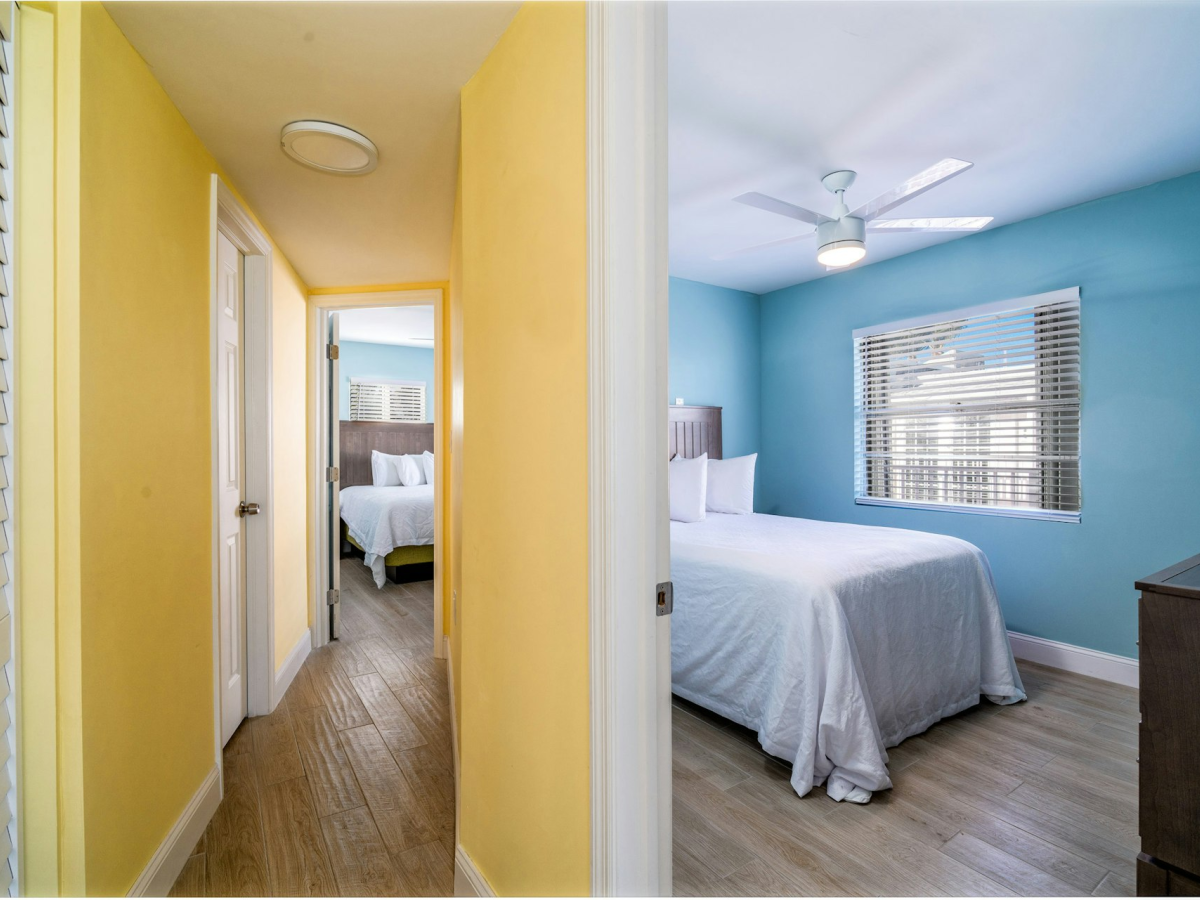 A bright bedroom with a bed, ceiling fan, blue walls, and window blinds, reached from a yellow hallway.