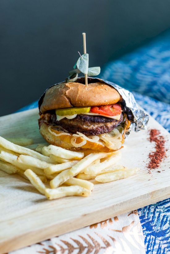 Cheeseburger with lettuce, tomato, pickles and fries on a wooden board, garnished with a toothpick herb.