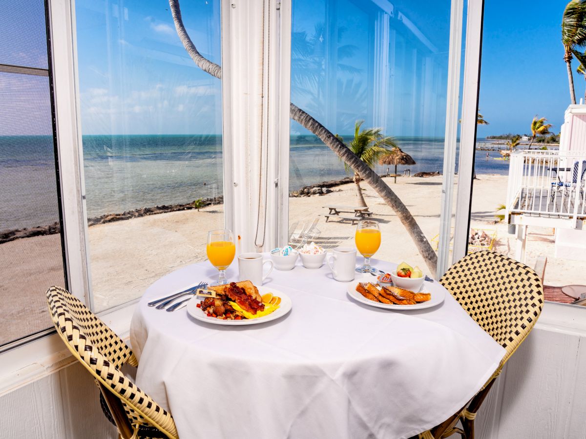 A small table set for two with plates of food and glasses of orange juice, by a window overlooking a beach with palm trees and the ocean.