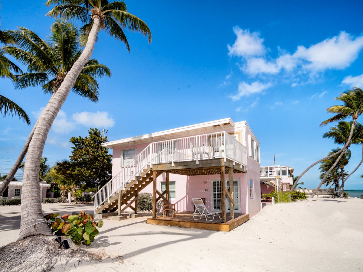 A pink beach house with a raised deck and surrounded by palm trees, situated on a sandy shore under a clear blue sky and calm ocean.