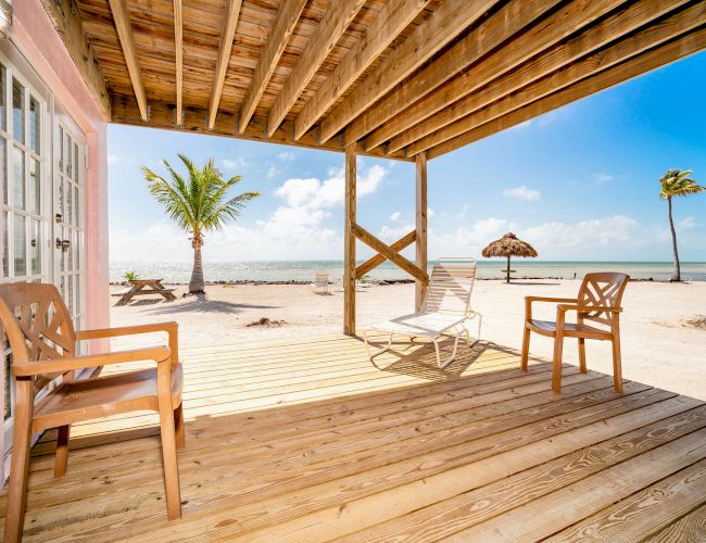 A wooden porch with chairs overlooks a sandy beach with palm trees, a picnic table, and a thatched umbrella under a bright blue sky.