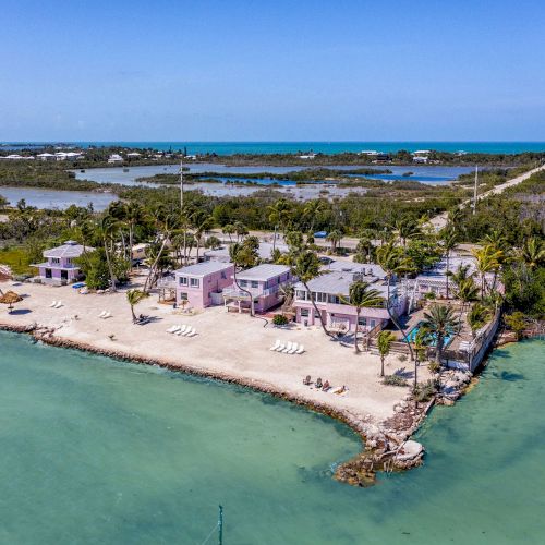 An aerial view of a coastal property with multiple buildings, palm trees, and lounge chairs on a sandy beach surrounded by turquoise waters.