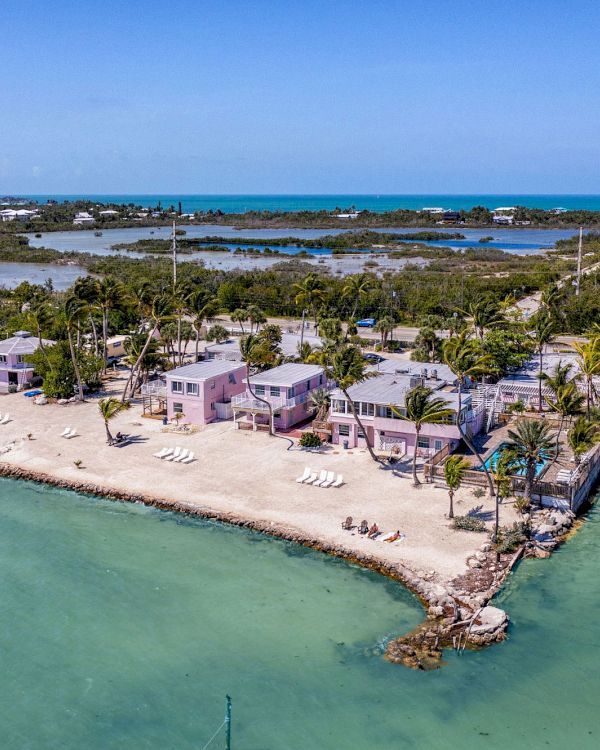 An aerial view of a coastal property with multiple buildings, palm trees, and lounge chairs on a sandy beach surrounded by turquoise waters.