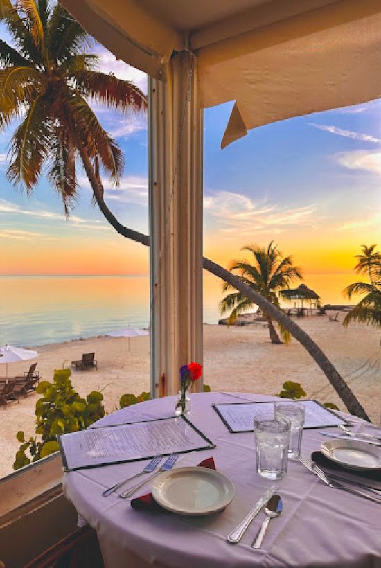 A seaside restaurant view with a set table, palm trees, and a sunset over the ocean, seen through large windows.