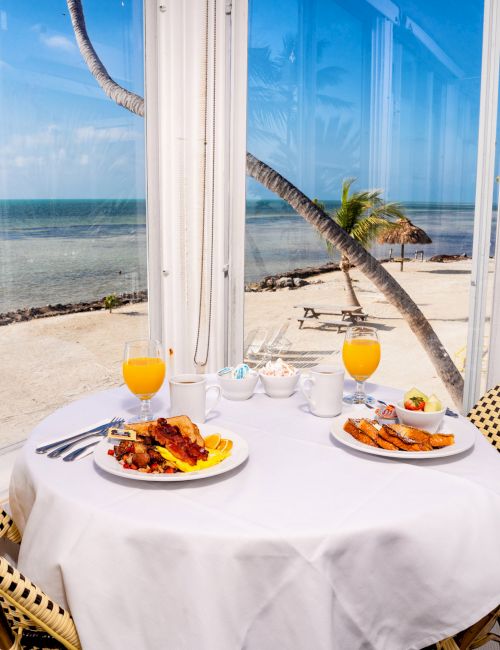 A beachfront table set for two with plates of breakfast food and glasses of orange juice, in a sunny, coastal dining area with an ocean view ending the sentence.